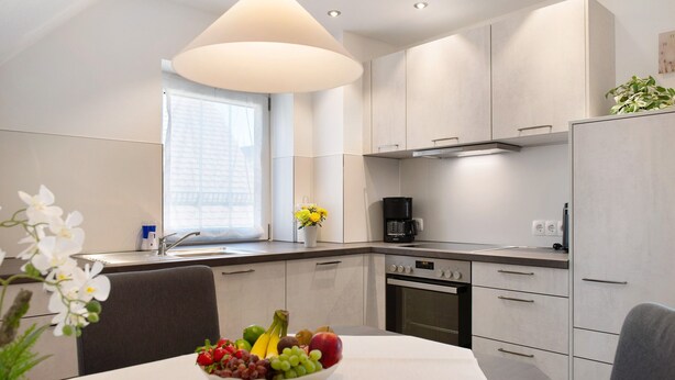 A modern kitchen with white cabinets, a dining table with a fruit bowl, and a window with a view. - Apartment 'Birnau' with Lake View and Wi-Fi (Hagnau)