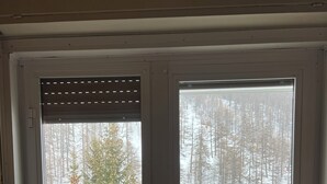 A room with a stone-tiled floor, a fireplace, a bed with a blanket, and two windows offering views of a snowy forest. - Carlotta whole apartment (Borgata Sestriere)