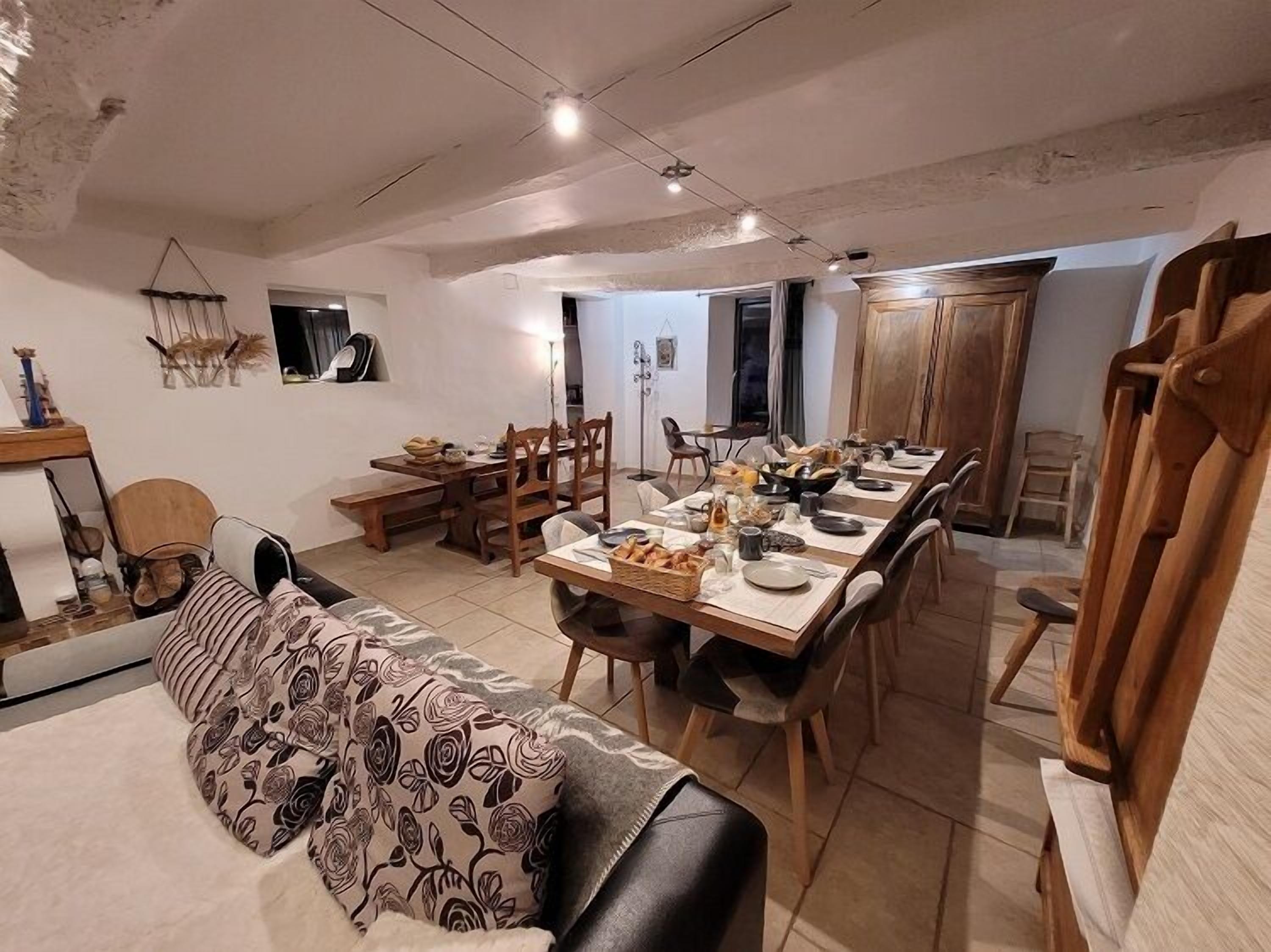 A dining area with a long wooden table set for a meal, surrounded by chairs. There is a sofa with a floral-patterned throw, a wooden bench, and a small table with decorative items. The room has a high ceiling with exposed beams and a wooden door.