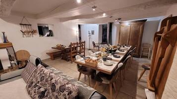 A dining area with a long wooden table set for a meal, surrounded by chairs. There is a sofa with a floral-patterned throw, a wooden bench, and a small table with decorative items. The room has a high ceiling with exposed beams and a wooden door.