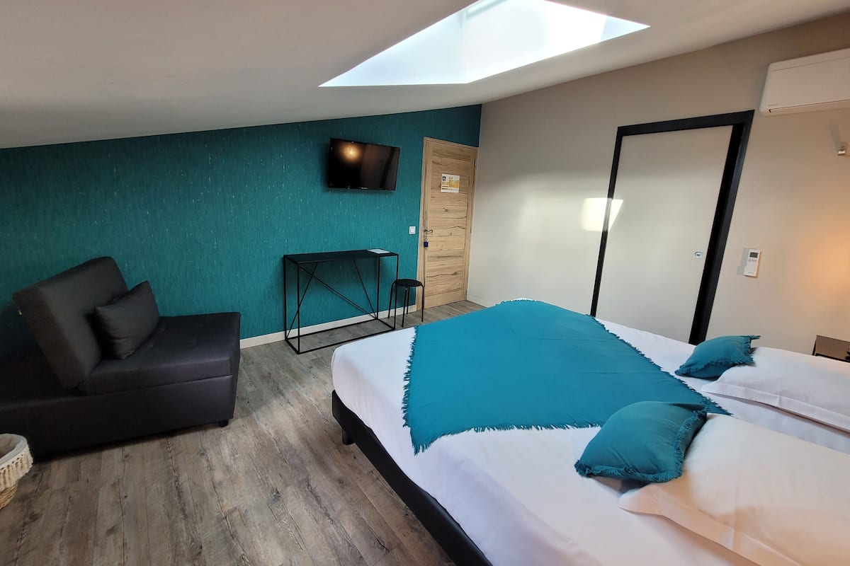 Une chambre moderne avec une lucarne, un téléviseur, un lit avec une couette turquoise, une petite table et un canapé.
