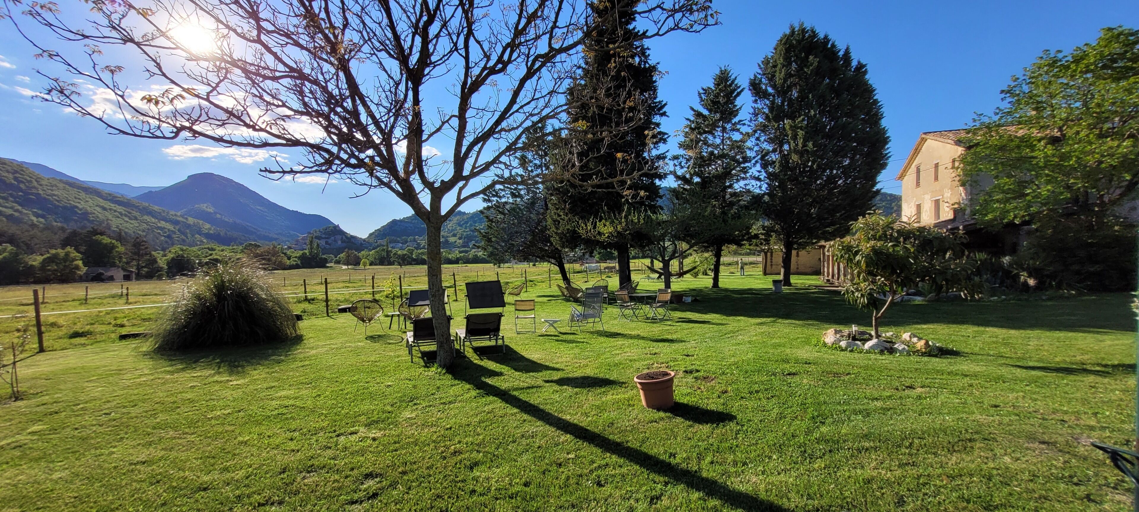 Un espacio al aire libre cubierto de césped, con algunas sillas y un pequeño árbol, rodeado de edificios y montañas.
