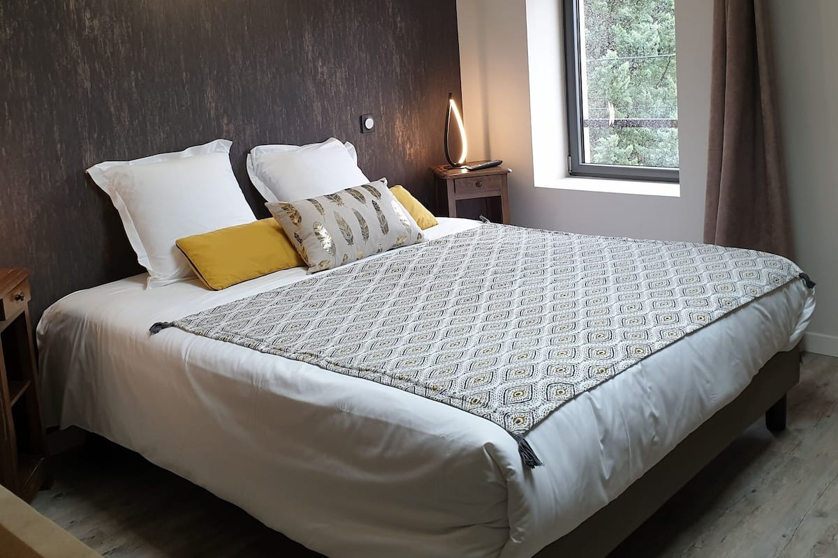 Une chambre avec un grand lit, une table de chevet, un climatiseur fixé au mur, une fenêtre avec des rideaux et une vue sur la verdure à l’extérieur.