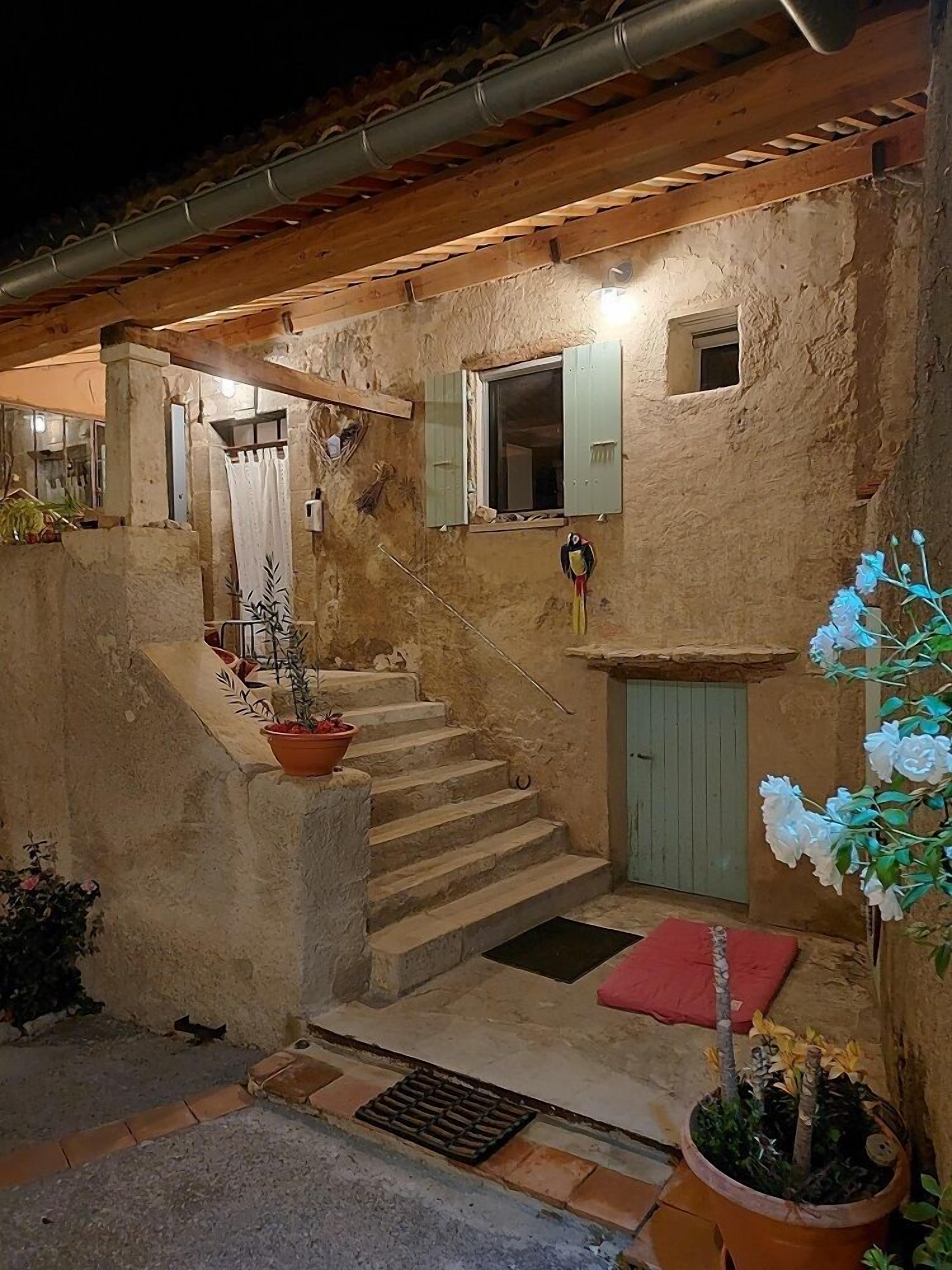 A rustic stone house with a wooden staircase leading to a door, a small window with shutters, and potted plants.