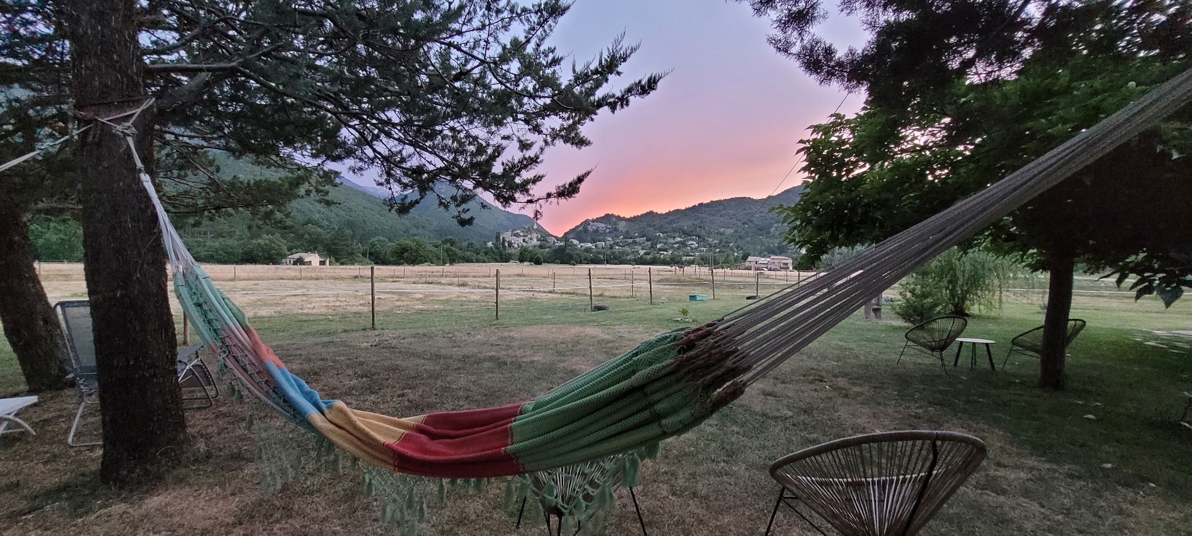 Un hamac est suspendu entre deux arbres dans une zone herbeuse, offrant une vue sur les montagnes et un coucher de soleil.