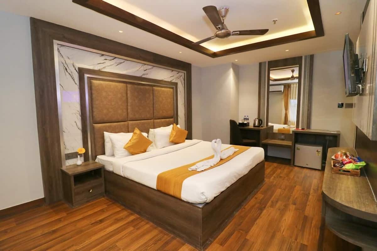 A hotel room with a large bed, wooden flooring, a ceiling fan, a small refrigerator, a bedside table with a lamp, and a large mirror.