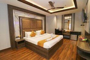 Une chambre d’hôtel avec un grand lit, un plancher en bois, un ventilateur de plafond, un petit réfrigérateur, une table de chevet avec une lampe et un grand miroir.