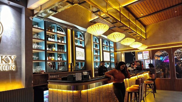 A bar area with shelves of bottles and glasses, a counter with a person standing, and a sign that reads "DRANK". - Transitel A Boutique Hotel (Barasat)