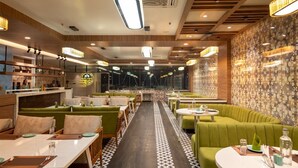 A restaurant with green booths and round tables, a patterned floor, and a bar area with shelves of bottles. - The Grand Royal Hotel (Samastipur)