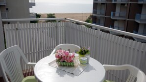 Ein Balkon mit rundem Tisch, zwei Stühlen, einer Blumenvase und einer Flasche. Der Balkon bietet Blick auf einen Strand und Gebäude.