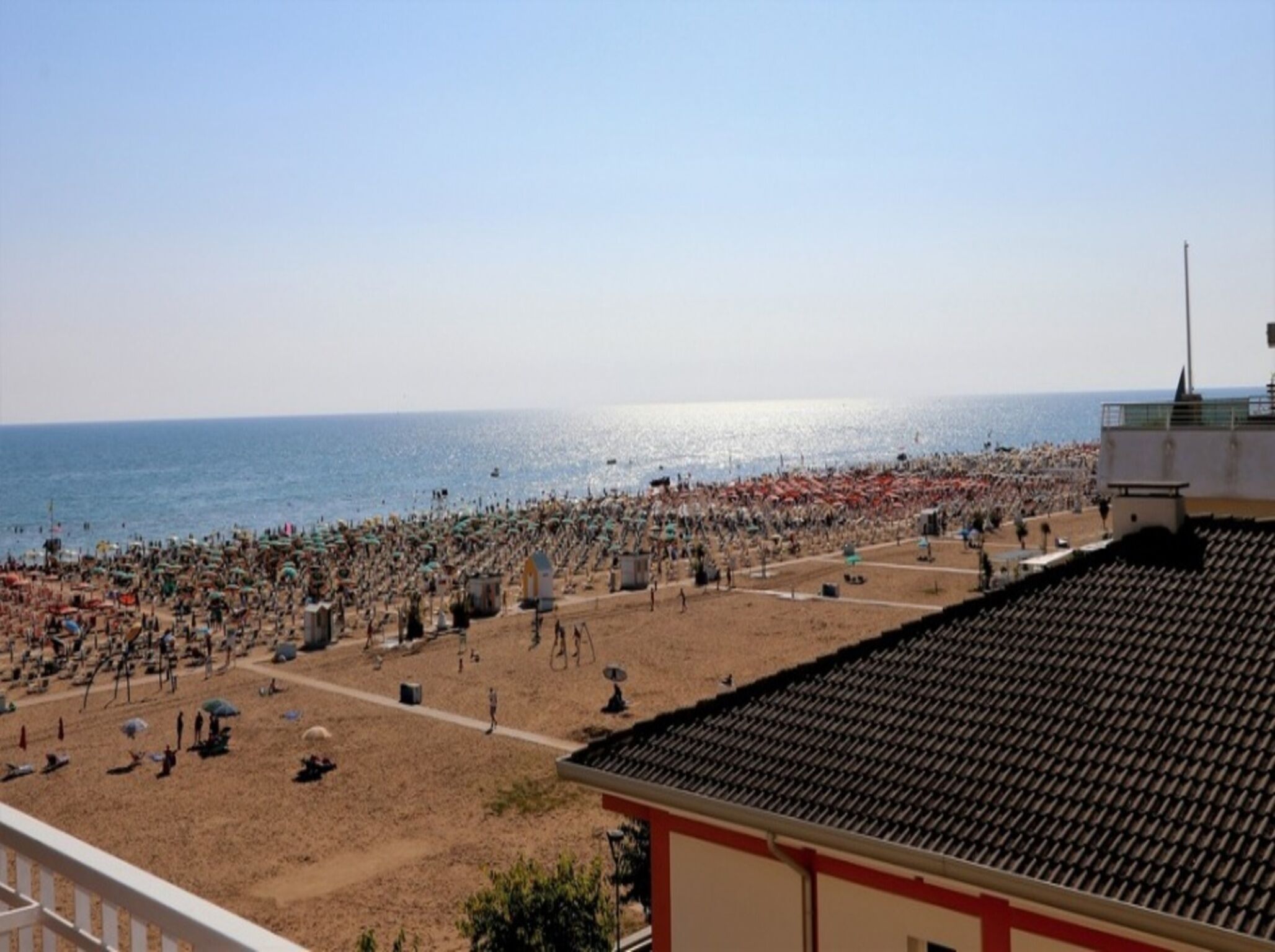 Una playa con numerosos sombrillas y gente, cielo despejado y un edificio con tejado de baldosas al frente.