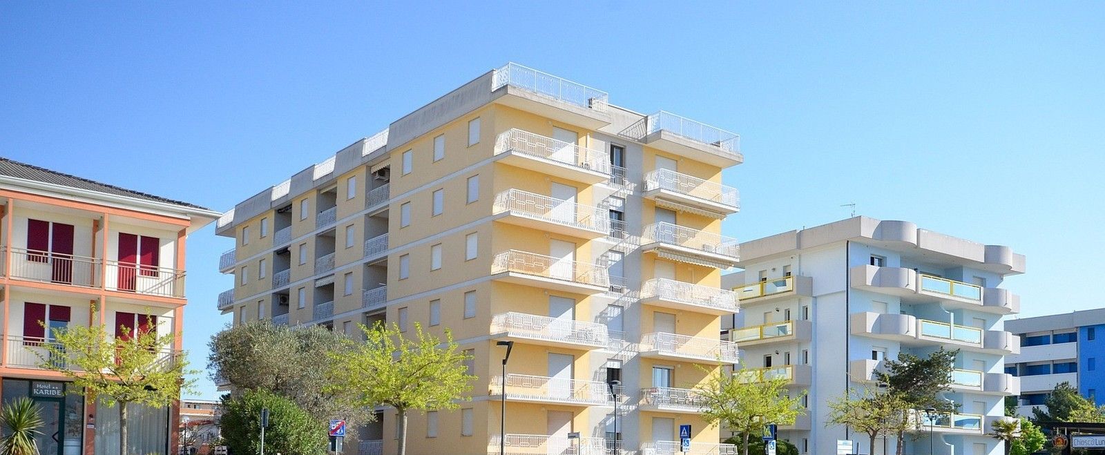 Un edificio residencial de varios pisos con balcones, cielo despejado y árboles al frente.