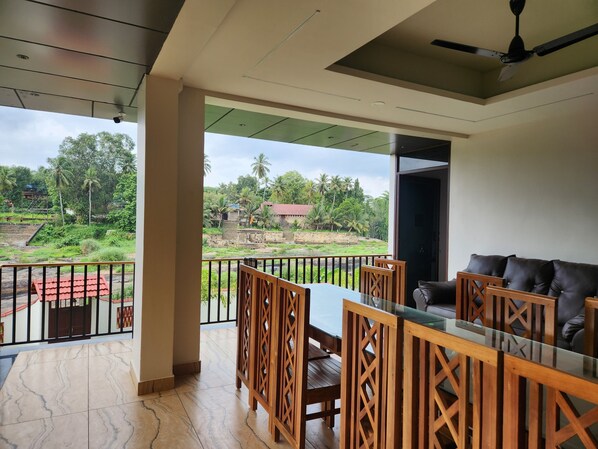 A balcony with a glass table and chairs, a black leather sofa, and a view of a green landscape with trees and a building. - Falls Regency (Vilavancode)