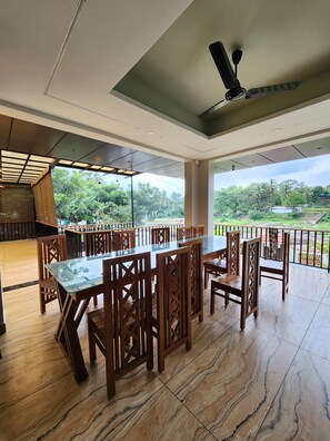 A spacious dining area with a glass table and wooden chairs, a ceiling fan, and a view of greenery outside. - Falls Regency (Vilavancode)