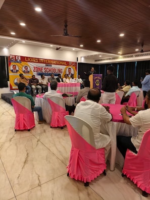 A conference room with round tables, attendees seated, and a banner on the wall. - Falls Regency (Vilavancode)