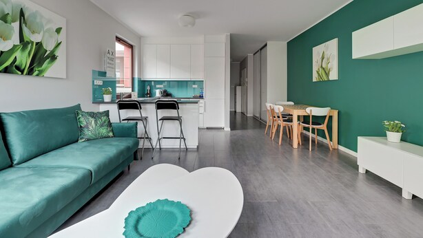 Un salon moderne avec un canapé de couleur turquoise, une table basse blanche et un coin cuisine doté de meubles blancs et d’un pare-feu turquoise.