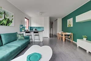 Una sala de estar moderna con un sofá verde azulado, una mesa de centro blanca y una cocina con armarios blancos y salpicadero verde azulado.