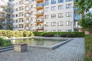 Un edificio residencial de varios pisos con balcones, un estanque pequeño y un paseo pavimentado.