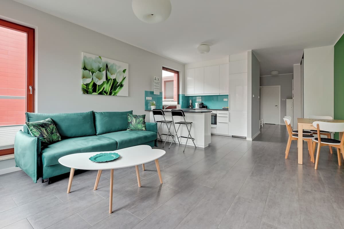 Una sala de estar moderna con un sofá verde, una mesa de centro blanca y una zona de cocina con armarios blancos y una pequeña isla.