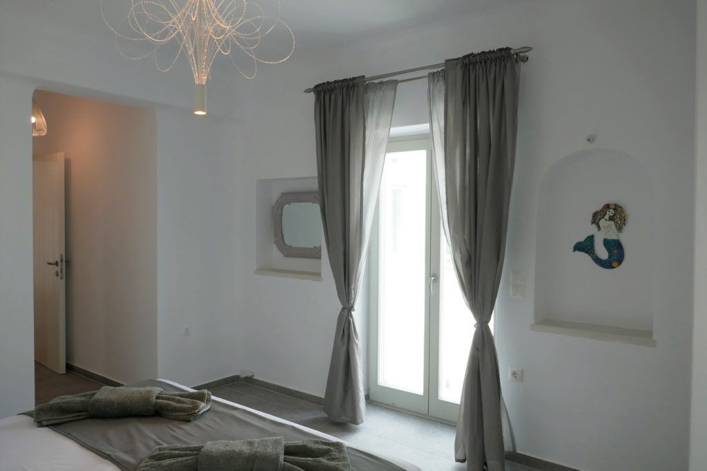 Un dormitorio con una cama, un ventanal con cortinas, una lámpara de pared y un estante pequeño con un objeto decorativo.