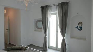 Un dormitorio con una cama, un ventanal con cortinas, una lámpara de pared y un estante pequeño con un objeto decorativo.