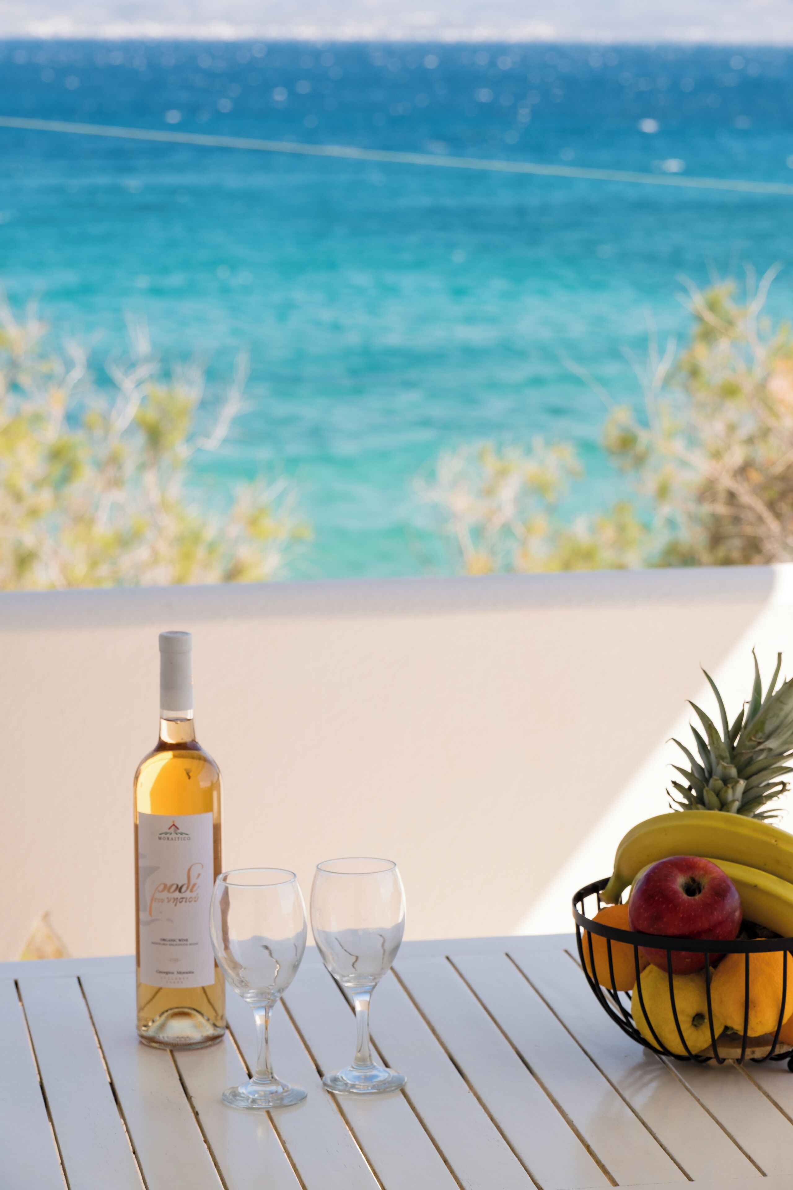 A table with a bottle of wine, two glasses, and a fruit basket by a seaside view.