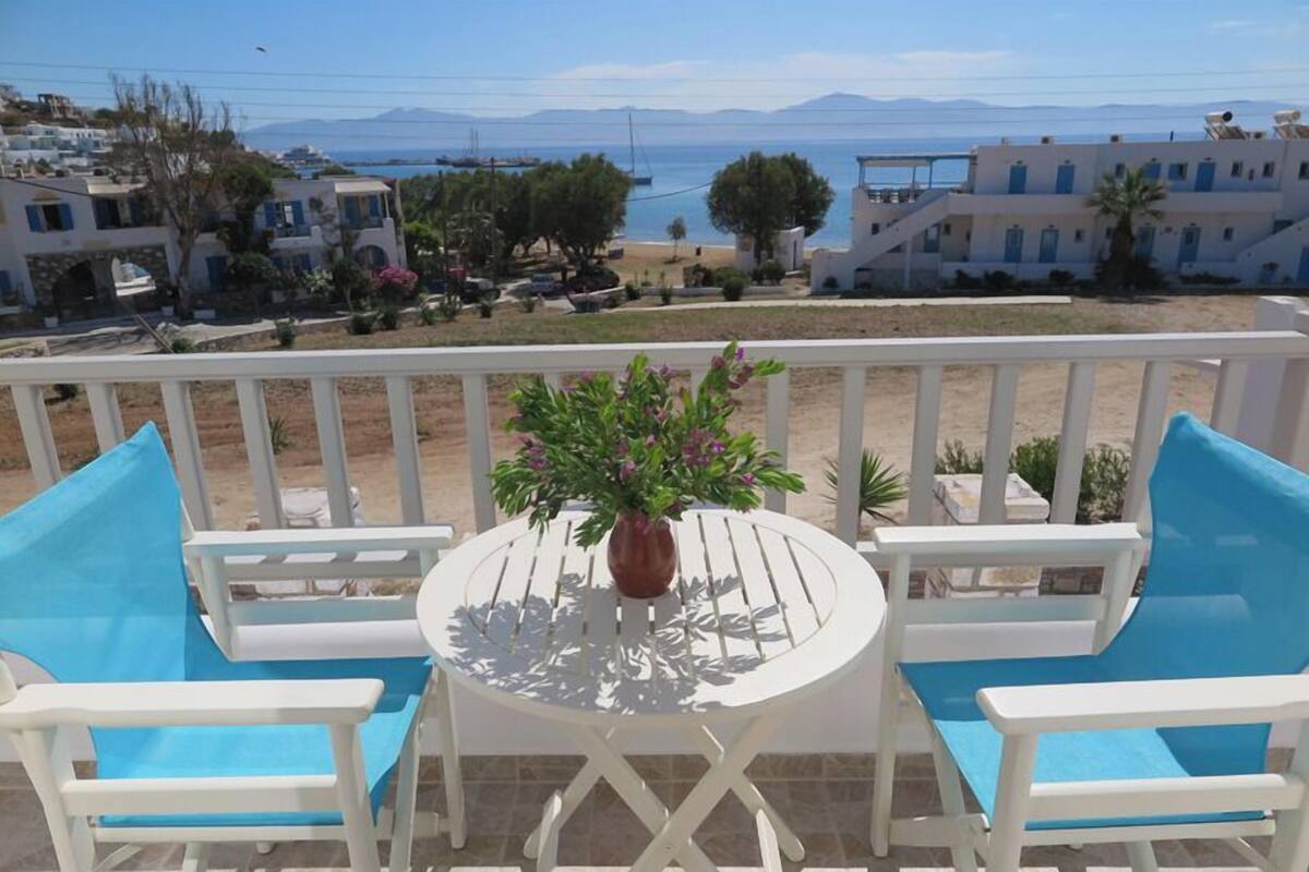 A balcony with white furniture and a potted plant, overlooking a beach and buildings.