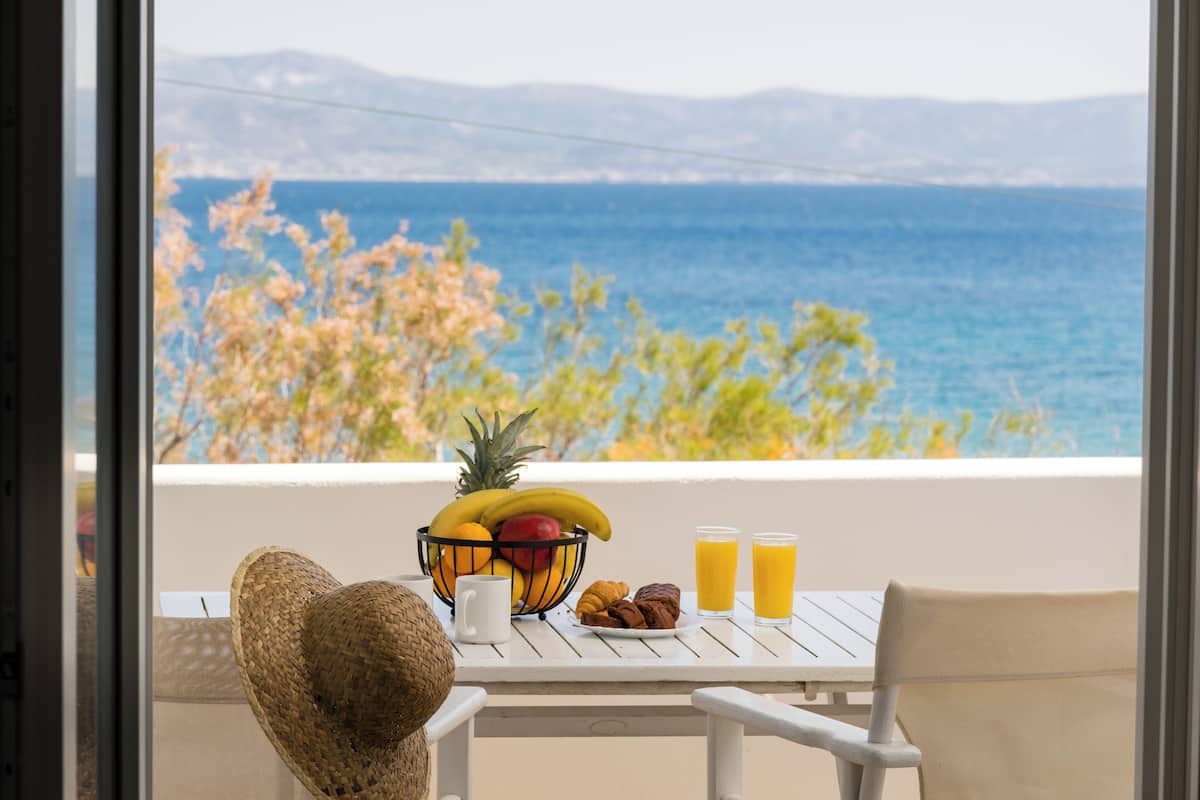 A table with a fruit bowl, croissants, and two glasses of orange juice, overlooking a scenic view of the sea and mountains.