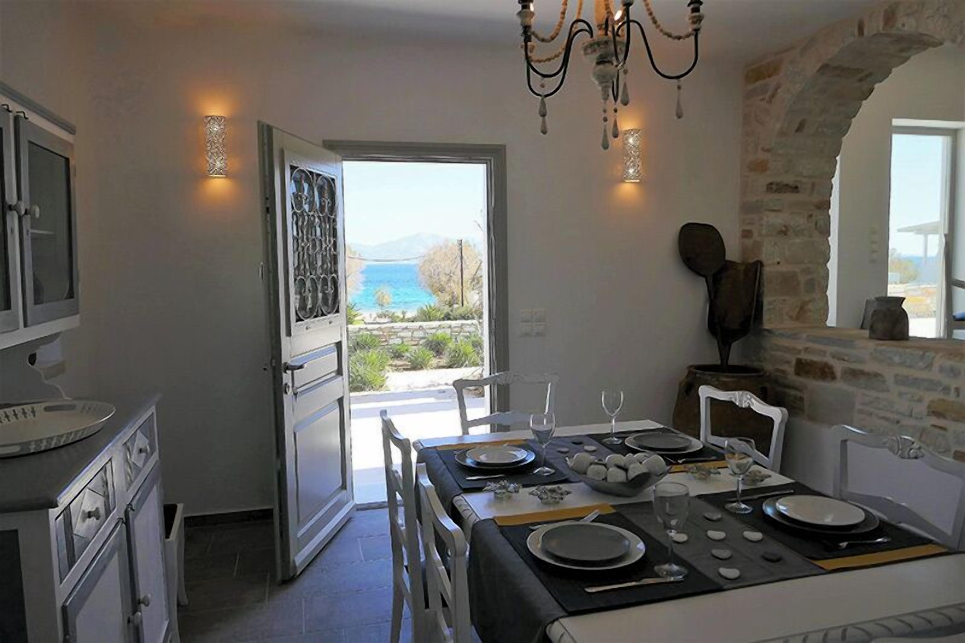 A dining area with a table set for four, a view of the sea through an open door, and a stone wall.