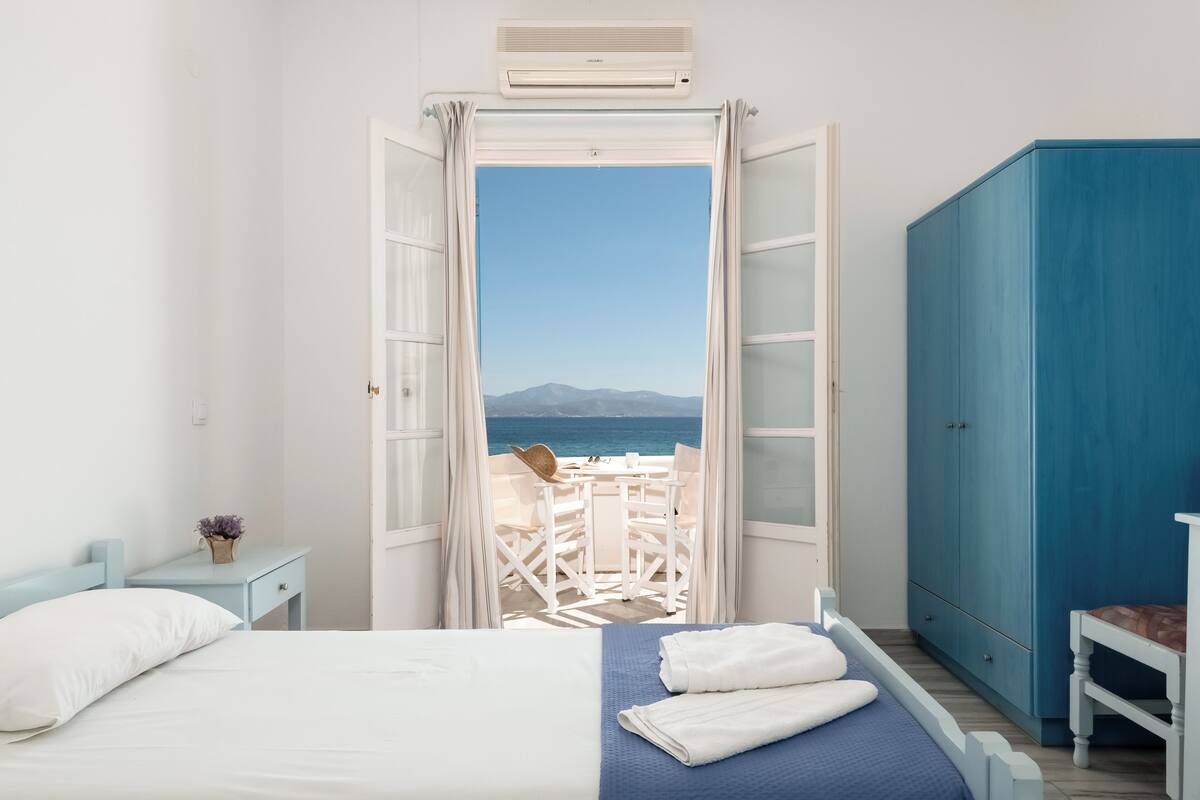 A bedroom with a bed, a nightstand, a wardrobe, and a view of the sea through an open door.