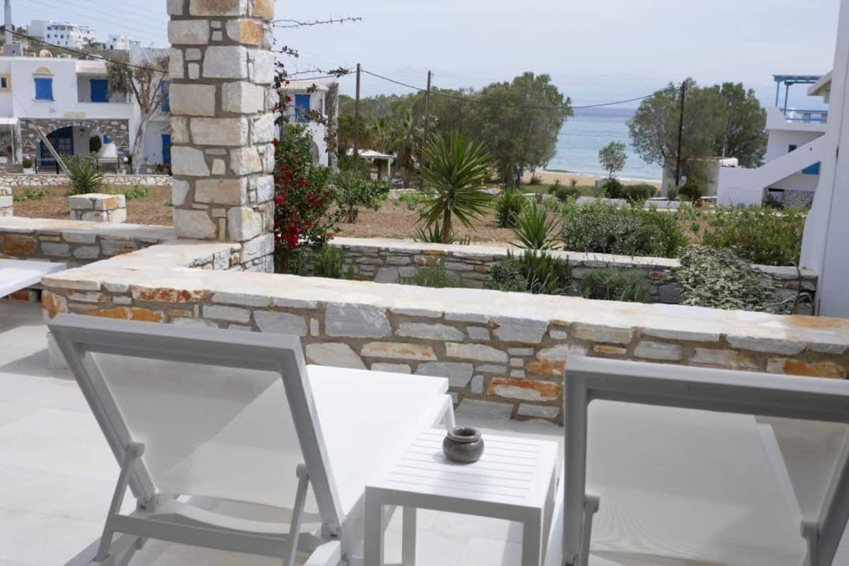 A terrace with white chairs and a stone wall, overlooking a beach and a village with white buildings.