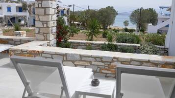 Una terraza con sillas blancas y un muro de piedra, con vista a una playa y un pueblo con edificios blancos.