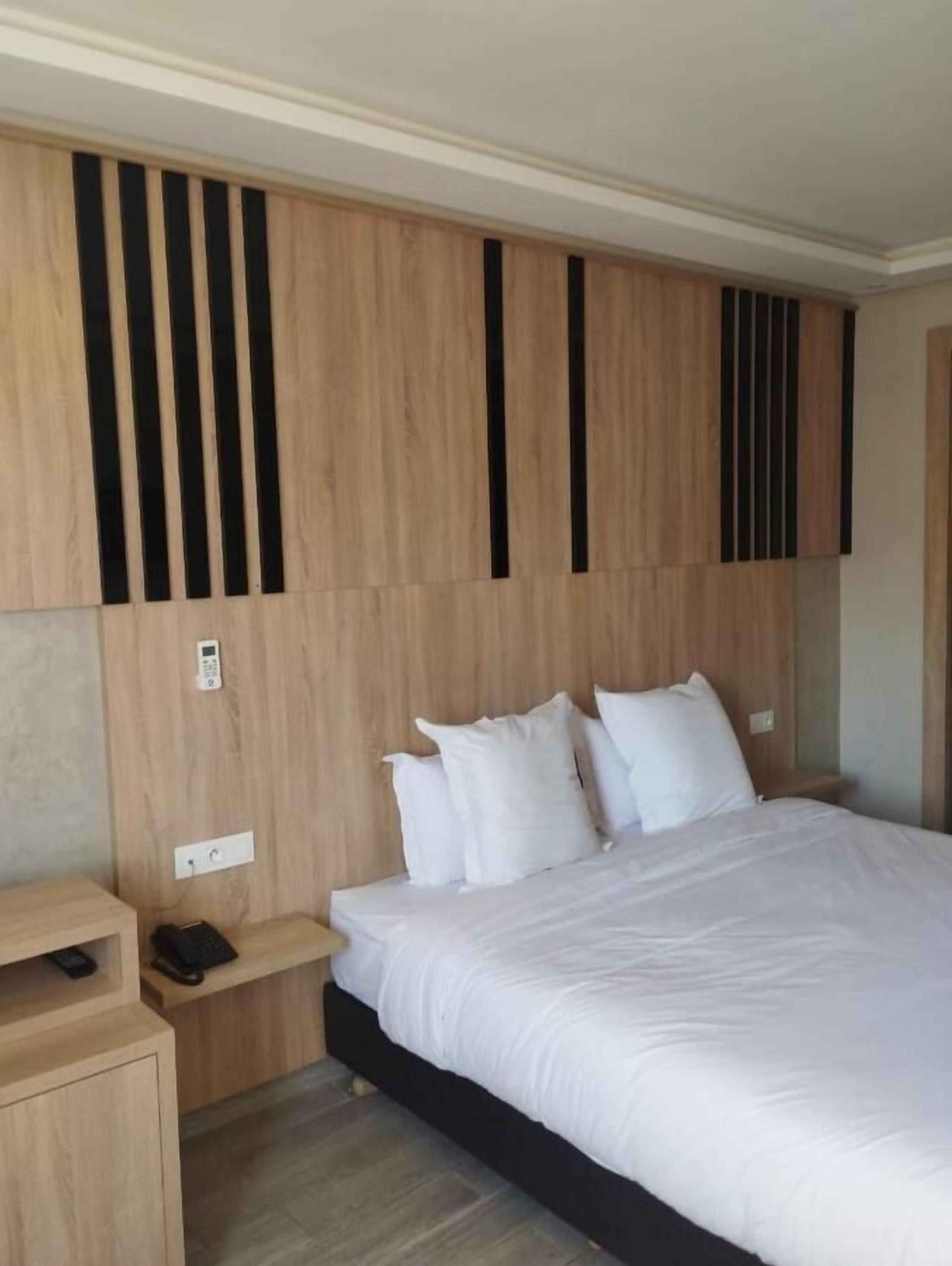 A modern hotel room with a large bed, wooden headboard with vertical black stripes, a bedside table with a phone, and a small wooden cabinet.