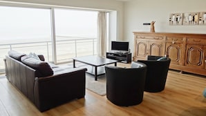 Ein modernes Wohnzimmer mit einer Couch, zwei Sesseln, einem Couchtisch und einem Fernseher. Ein großes Fenster bietet Blick aufs Meer.