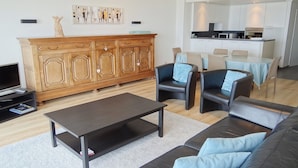 Ein modernes Wohnzimmer mit einem dunklen Couchtisch, Ledersofas und einem Holzkabinett. Ein Fernseher steht auf einem Lowboard links, und im Hintergrund ist ein Essbereich mit Stühlen und einem Tisch zu sehen.
