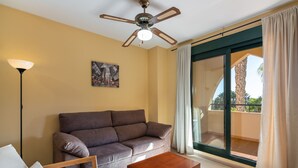 Un salon comprenant un canapé, une table basse, un lampadaire, un ventilateur de plafond, un tableau et une porte-fenêtre coulissante donnant sur un balcon.