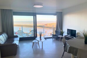 A modern living room with a large window, a sofa, a coffee table, a TV, and a dining area. - Studio in Nieuwpoort with Sea View Terrace (Nieuwpoort)