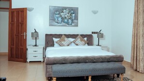 A bedroom with a bed, two bedside tables with lamps, a wooden door, a painting on the wall, and a patterned curtain. - TB 11 (Kibaoni)