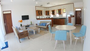 A modern living room with a sofa, coffee table, and chairs, a kitchen with wooden cabinets and a refrigerator, and a flat-screen TV mounted on the wall. - TB 11 (Kibaoni)