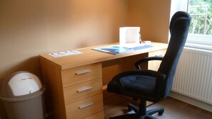 A small office space with a wooden desk, a black office chair, a waste bin, and a window with a view of greenery. - Vacation apartment Luna (Aurich)
