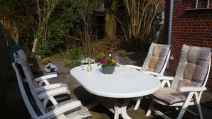 A round outdoor table with four white chairs, surrounded by greenery and a brick wall. - Vacation apartment Luna (Aurich)