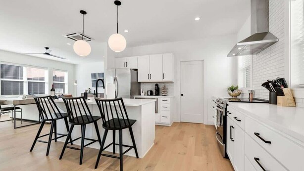 Una cocina moderna con armarios blancos, una isla central con taburetes, un extractor de acero inoxidable y vistas de la ciudad a través de amplias ventanas.