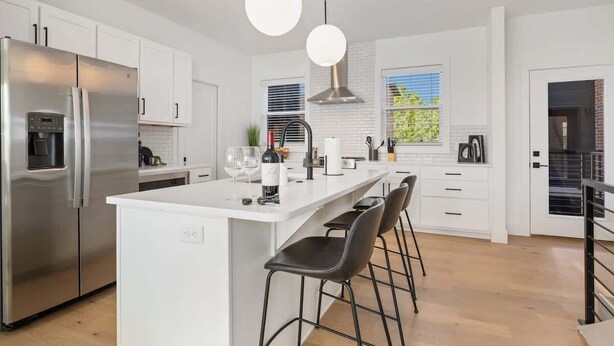 Une cuisine moderne avec des meubles blancs, un réfrigérateur en acier inoxydable, un îlot central doté de tabourets de bar et un éclairage suspendu.