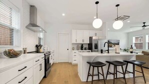 Cozinha moderna com armários brancos, uma ilha central com banquetas e uma grande janela com persianas.