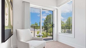 A room with a white armchair, a small round table, a large sliding glass door with a view of greenery, and a painting on the wall.