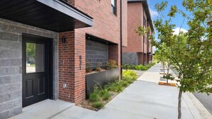 A modern brick building with a black door, a small garden area with plants, and a tree on the sidewalk.