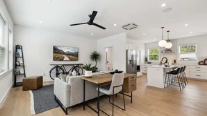 A modern kitchen with a central island, a dining area with a sofa and a desk, and a bicycle in the background.