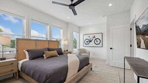 A bedroom with a large bed, a ceiling fan, a bicycle artwork on the wall, and a view of the outdoors through the windows.