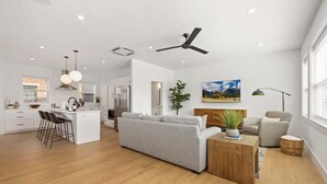 Una cocina moderna con armarios blancos, una isla central con taburetes de bar, un sofá gris y una mesa de centro de madera.