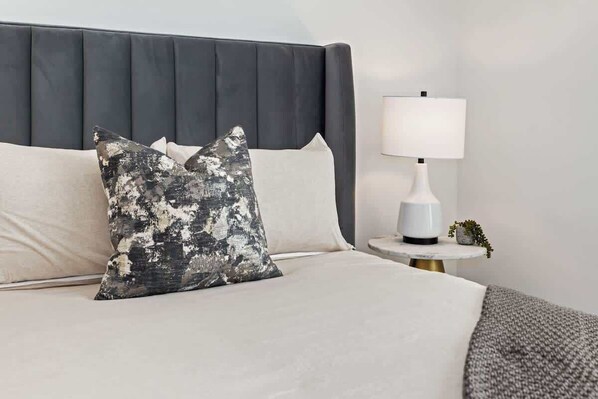 Een keurig opgemaakt bed met een grijs bekleed hoofdbord, een witte nachtkastje met een lamp en een kleine potplant.